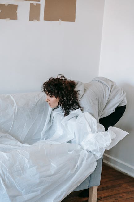 A woman with curly dark hair, dressed in a light grey hoodie and black trousers, is leaning over and adjusting or covering a white sofa with a moving blanket or protective wrapping material inside a well-lit room. The sofa is positioned against a white wall, which has a few unpainted patches or sections covered with tape, indicating ongoing renovation or preparation for a house removal. The flooring is dark wood, and the scene appears to be part of a home relocation or furniture transport process, as managed by Man and Van Isleworth, which specializes in removals. The woman is engaged in securing or preparing furniture for moving, emphasizing careful handling during packing and loading stages.