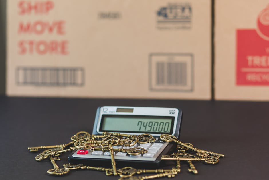 A digital calculator displaying the number 749,800 is placed on a black surface, surrounded by numerous antique brass keys of various shapes and sizes, some with ornate handles. In the background, blurry cardboard moving boxes with red and black printed labels are stacked, indicating packaging for a home relocation. The scene suggests packing and inventory processes involved in furniture transport and house removals, with the keys possibly symbolising security or ownership during the moving process. The environment appears to be a loading or preparation area, illuminated with neutral lighting, emphasizing the tools and materials used for organized moving services. Man and Van Isleworth's removals service collaborates with such logistics for efficient house moves, as indicated in the related content.