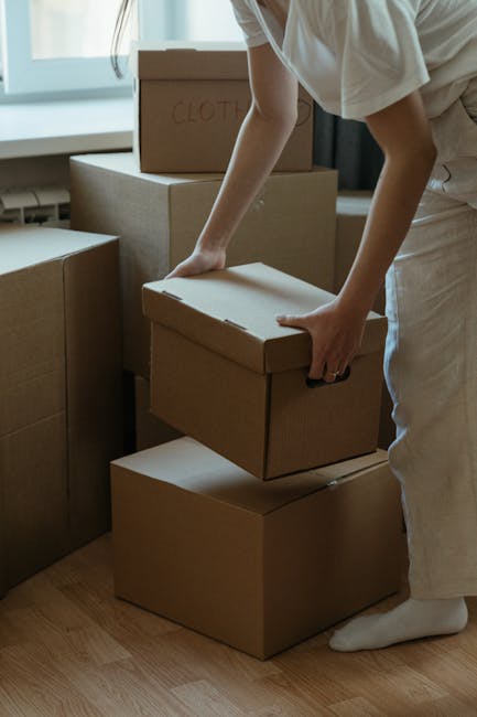 A woman with curly dark hair, dressed in a light grey hoodie and black trousers, is leaning over and adjusting or covering a white sofa with a moving blanket or protective wrapping material inside a well-lit room. The sofa is positioned against a white wall, which has a few unpainted patches or sections covered with tape, indicating ongoing renovation or preparation for a house removal. The flooring is dark wood, and the scene appears to be part of a home relocation or furniture transport process, as managed by Man and Van Isleworth, which specializes in removals. The woman is engaged in securing or preparing furniture for moving, emphasizing careful handling during packing and loading stages.