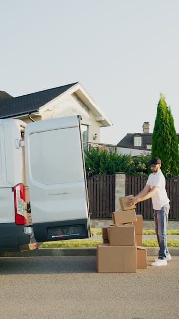 A man wearing casual clothing is standing on the street next to an open white delivery van with its rear door raised, engaged in a home relocation process. He is handling cardboard boxes of various sizes, stacking some on the pavement while others are ready to be loaded or unloaded. The boxes are made of brown corrugated cardboard, and a few are sealed with packing tape. The van is parked close to a residential driveway, with a small garden featuring green bushes and a tall, slim conifer tree behind a dark wooden fence. In the background, there is a modern house with a pitched roof and a chimney, under a clear pale blue sky. The scene captures the typical loading or unloading activities involved in furniture transport and packing during a move, with the focus on the careful handling of packing materials as part of the removal process undertaken by Man and Van Isleworth.