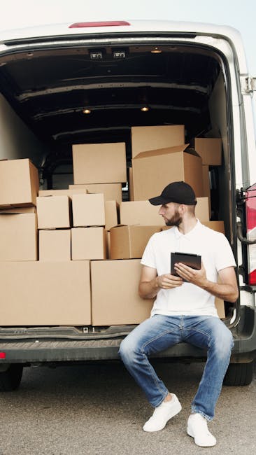 A man wearing a white t-shirt, blue jeans, white sneakers, and a black cap is seated on the pavement beside an open delivery van during a home relocation. The van's rear doors are wide open, revealing a cargo space filled with numerous brown cardboard boxes of various sizes, stacked neatly to maximise space. Some boxes are wrapped in transparent plastic film, indicating packed or protected household items. The man is holding a tablet or mobile device, possibly coordinating the packing and loading process. Inside the cargo area, the boxes are arranged on the floor and on top of each other, with additional packing materials such as small boxes and packing paper visible. The environment is outdoors, with the van parked on the street close to an entrance of a house or building, suggesting an ongoing furniture transport and moving logistics operation. This scene exemplifies careful packing and loading procedures in residential removals, illustrating services provided by Man and Van Isleworth as part of a professional removals operation.