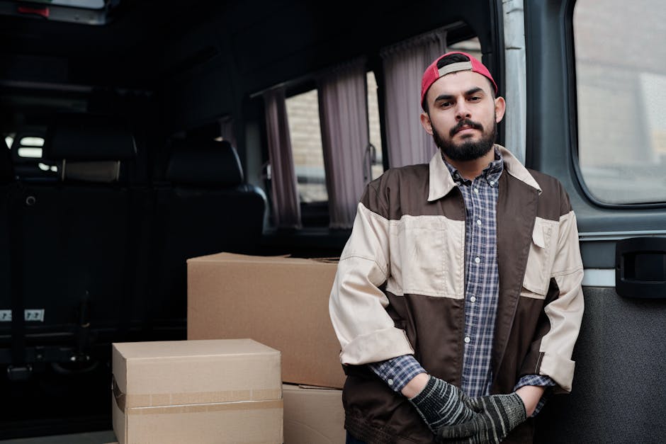 A man wearing casual clothing is standing on the street next to an open white delivery van with its rear door raised, engaged in a home relocation process. He is handling cardboard boxes of various sizes, stacking some on the pavement while others are ready to be loaded or unloaded. The boxes are made of brown corrugated cardboard, and a few are sealed with packing tape. The van is parked close to a residential driveway, with a small garden featuring green bushes and a tall, slim conifer tree behind a dark wooden fence. In the background, there is a modern house with a pitched roof and a chimney, under a clear pale blue sky. The scene captures the typical loading or unloading activities involved in furniture transport and packing during a move, with the focus on the careful handling of packing materials as part of the removal process undertaken by Man and Van Isleworth.