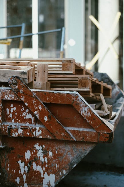A close-up view of a rusty, weathered boat used for home relocation or transport, loaded with multiple light-colored wooden pallets and crates stacked one above the other inside the vessel near the bow. The pallets are constructed from smooth wood with slatted designs, secured with metal nails, and some are wrapped with plastic or fabric for protection. The boat is positioned on pavement or a dock area outside a building with large glass windows, reflecting the surrounding environment and natural light. In the background, there are blurred structures, possibly scaffolding or construction equipment, indicating an active site. The boat's surface shows white paint splatters and signs of corrosion, emphasizing its outdoor, utilitarian purpose. This scene exemplifies the logistics involved in furniture transport and packing during a house move, and it aligns with the services provided by Man and Van Isleworth for efficient removals and moving logistics.