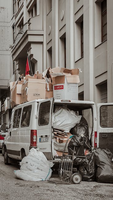 A white van parked on a city street with its rear doors open, revealing a large collection of packed cardboard boxes, black plastic garbage bags, and wrapped furniture materials inside, ready for loading during a house relocation process. Additional boxes and materials are stacked on the van's roof, secured with straps, with some boxes labelled, indicating packing for a move. The van is positioned close to a multi-storey building with a grey facade, and a small trolley is seen on the pavement nearby, supporting the loading activities. This scene illustrates the transport phase of home removals, with visible packing supplies, trolleys, and the van prepared for furniture transport, typical of professional moving services such as those offered by Man and Van Isleworth, aligning with local rules for waste disposal during moves as referenced on the Isleworth council guidelines page.
