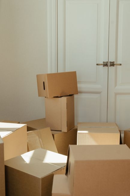 A collection of unpacked cardboard moving boxes in various sizes, some stacked and others placed on the floor, inside a room with cream walls and a closed white door with a brass handle. The boxes are