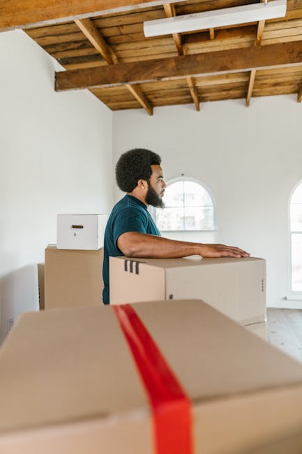 A man with dark curly hair and a beard, wearing a blue t-shirt, is inside a bright, minimally decorated room with white walls and large arched windows. He is standing behind several medium-sized cardboard boxes, one of which features a red packing strap, indicating they are being prepared for a move. The ceiling above has an exposed wooden beam structure, adding a natural, rustic element to the space. The room receives ample natural light from the windows, highlighting the packing and loading process typical of house removals. Nearby, additional boxes of various sizes are visible, suggesting organized packing for a home relocation. This scene reflects the loading phase of furniture transport and packing associated with professional removals services, such as those offered by Man and Van Isleworth, demonstrating careful handling of household items before loading them into a van for transportation. The environment and objects emphasize the preparation and logistics involved in house move operations, aligning with the focus on removals, relocation, and safety.