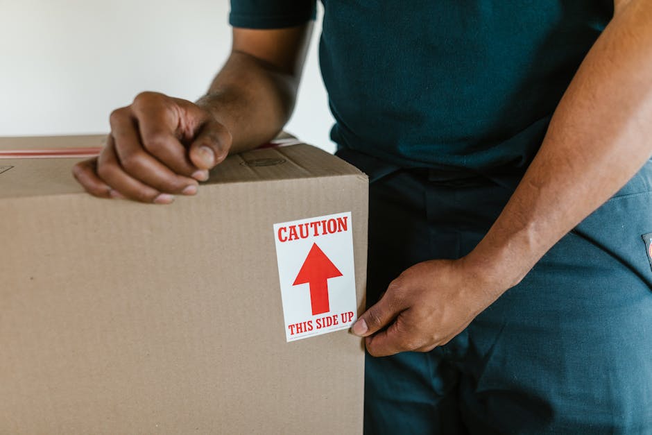 A man with dark curly hair and a beard, wearing a blue t-shirt, is inside a bright, minimally decorated room with white walls and large arched windows. He is standing behind several medium-sized cardboard boxes, one of which features a red packing strap, indicating they are being prepared for a move. The ceiling above has an exposed wooden beam structure, adding a natural, rustic element to the space. The room receives ample natural light from the windows, highlighting the packing and loading process typical of house removals. Nearby, additional boxes of various sizes are visible, suggesting organized packing for a home relocation. This scene reflects the loading phase of furniture transport and packing associated with professional removals services, such as those offered by Man and Van Isleworth, demonstrating careful handling of household items before loading them into a van for transportation. The environment and objects emphasize the preparation and logistics involved in house move operations, aligning with the focus on removals, relocation, and safety.
