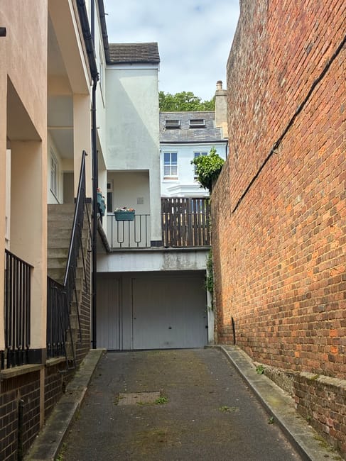 A narrow residential driveway situated between a multi-story white building on the left and a tall red brick wall on the right. The driveway slopes gently downward and is paved with asphalt, with some patches of moss or dirt visible. At the top of the driveway, there is a small balcony with black metal railings, beneath which a closed garage door is paint in light grey. In the background, a white building with multiple windows, greenery including a tree, and a grey roof are visible. The scene is well-lit by natural daylight, suggesting a clear day. This setting is typical for house removals and furniture transport logistics, with available access to the upper-level balcony and garage for loading or unloading. Man and Van Isleworth is prepared to manage such tight-access relocations, offering suitable equipment like trolleys and straps for safe furniture handling.