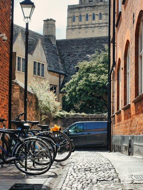 A narrow cobblestone street in Isleworth, shown during daytime, lined with red brick and stone buildings on either side. On the left, several bicycles are parked against a black metal bike rack, some secured with locks, with a nearby black street lamp. In the background, a blue van is parked close to a stone wall with greenery and flowering trees behind it. The scene suggests a typical residential area with limited parking access, consistent with house removals and local relocation services. The image is well-lit by natural daylight, capturing the tight urban environment where careful loading and parking solutions are necessary for moving and furniture transport. It appears to be part of a professional removal service, such as Man and Van Isleworth, providing access coordination in this historic street of Isleworth.