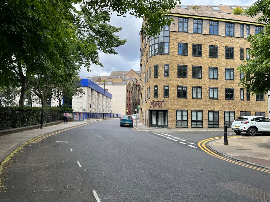 A street scene during a home relocation in Isleworth, showing a curved road with a parked white car and a dark blue car near the curb, and a man and van from Man and Van Isleworth parked on the pavement close to a large, multi-story brick building with numerous black-framed windows. The building has a rounded corner facing the street, with a small entrance door at ground level. On the left side, there is a park area with lush green trees and a black metal fence, partially shading the sidewalk where a person is walking. Behind the building, a modern white construction or retail structure with blue accents and decorative textures on its facade is visible, along with other nearby buildings. The scene is illuminated by daylight with a mix of sun and clouds, and the area appears to be in the process of furniture transport or packing as part of an office move, supported by the presence of the vehicle and the urban environment.