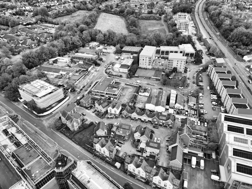 Aerial view of a riverside town showing a commercial street with multiple buildings, cars parked along the road, and a small parking area adjacent to the water. The scene includes a river with several boats docked along the quay, as well as green spaces and trees lining the waterfront. The photograph captures a clear day with natural lighting, highlighting the urban layout and surrounding residential areas. This image relates to house and office removals, illustrating logistical considerations for transport and relocation services provided by Man and Van Isleworth as part of their property moving and furniture transport solutions near Syon Lane in Isleworth.