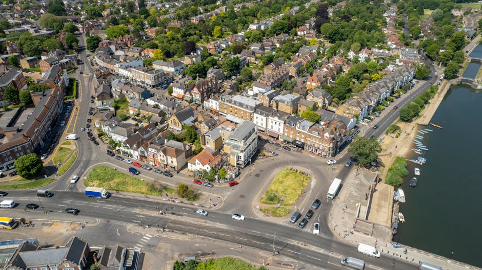Aerial view of a town centre in Isleworth with a mix of residential and commercial buildings, including shops and apartments, situated along a river with a small marina. The image shows a busy street with multiple lanes of traffic, including cars, trucks, and vans, some of which are likely part of a home removal or moving logistics operation. Near the waterfront, there is a paved area with parked vehicles and a few small boats docked at the marina, indicating relocation services involving navigation or transportation by water. The surrounding streets are lined with trees, and the properties vary in architectural style, with some older brick buildings and more modern structures. Inside the town, there are visible green spaces, pedestrian pathways, and parking areas, all captured under bright daylight, supporting efficient packing and moving processes by companies such as Man and Van Isleworth.
