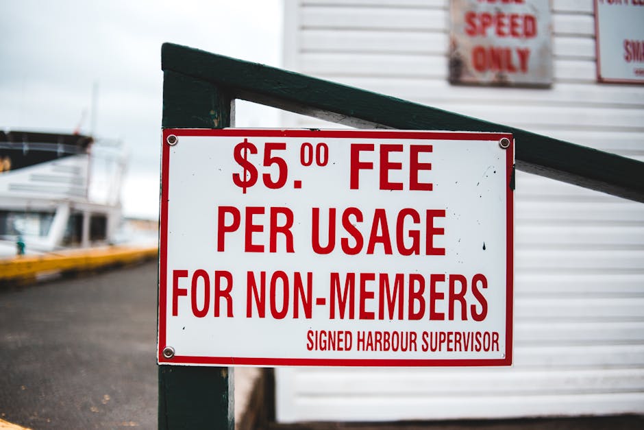 Close-up of a rectangular white sign with red text displaying a fee structure, reading '$5.00 FEE PER USAGE FOR NON-MEMBERS' in large letters, with a smaller line at the bottom stating 'SIGNED HARBOUR SUPERVISOR.' The sign is mounted on a dark green metal frame attached to a wooden post. The background shows part of a building with a white exterior wall and a partially visible red and white sign mounted above, along with a paved area and some boats docked on the water, indicating a harbour or marina setting. The scene is outdoors with natural lighting, capturing the context of areas where house removals may occur near marinas or docks, emphasizing logistical considerations for transport or storage outside residential or commercial properties, as typical in house moves or furniture transport services.
