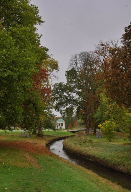 A wide view of Osterley Park in bright daylight shows a large, well-maintained lawn with a decorative pink and green emblem at its centre, bordered by symmetrical gravel pathways. Several neatly trimm