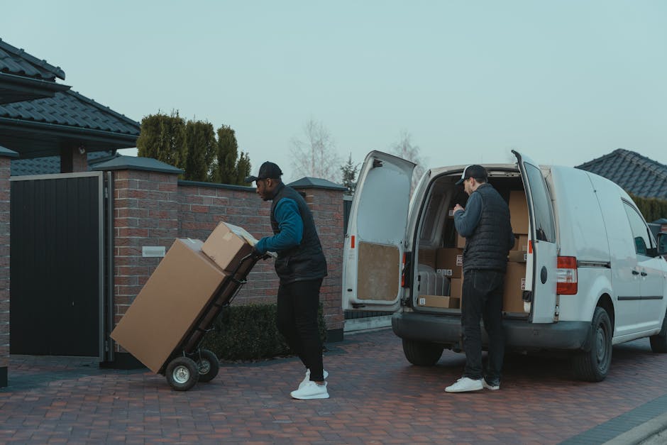 Two men engaged in a home relocation process outside a residential property, loading cardboard boxes and furniture onto a white van as part of a furniture transport and packing and moving service. The first man, wearing a black cap, glasses, a dark vest over a blue long-sleeve shirt, and white sneakers, is pushing a hand truck loaded with a large cardboard box towards the back of the van, which has its rear doors open revealing additional packed boxes inside. The second man, dressed in a black baseball cap, dark jacket, and black pants with white sneakers, is standing near the van's open side door, organizing or preparing other boxes inside. The setting is an outdoor driveway, paved with brick, adjacent to a brick boundary wall with a black gate, under overcast sky, with residential houses visible in the background. This scene depicts the logistics involved in packing, loading, and transporting household items during a home removal, with equipment such as trolleys and cardboard boxes being used by Man and Van Isleworth, supporting efficient moving services.