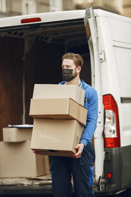 A man wearing a black face mask and a blue jacket is standing outside the open rear doors of a white moving van, holding three cardboard boxes of varying sizes, with one box balanced on top of the others. Inside the van, several additional cardboard boxes and packing materials are visible, some stacked on the floor. The scene appears to be part of a home relocation or furniture transport process, with the man preparing to load or unload the boxes during packing and moving activities. The background shows a blurred cityscape, suggesting an urban environment, and the lighting indicates daytime. This image exemplifies the packing, loading, and logistical aspects of professional removals services provided by Man and Van Isleworth, supporting efficient house removals or moving logistics.