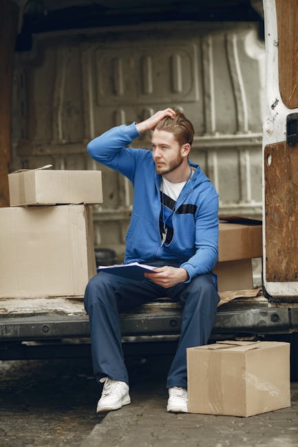 Two men engaged in a home relocation process outside a residential property, loading cardboard boxes and furniture onto a white van as part of a furniture transport and packing and moving service. The first man, wearing a black cap, glasses, a dark vest over a blue long-sleeve shirt, and white sneakers, is pushing a hand truck loaded with a large cardboard box towards the back of the van, which has its rear doors open revealing additional packed boxes inside. The second man, dressed in a black baseball cap, dark jacket, and black pants with white sneakers, is standing near the van's open side door, organizing or preparing other boxes inside. The setting is an outdoor driveway, paved with brick, adjacent to a brick boundary wall with a black gate, under overcast sky, with residential houses visible in the background. This scene depicts the logistics involved in packing, loading, and transporting household items during a home removal, with equipment such as trolleys and cardboard boxes being used by Man and Van Isleworth, supporting efficient moving services.