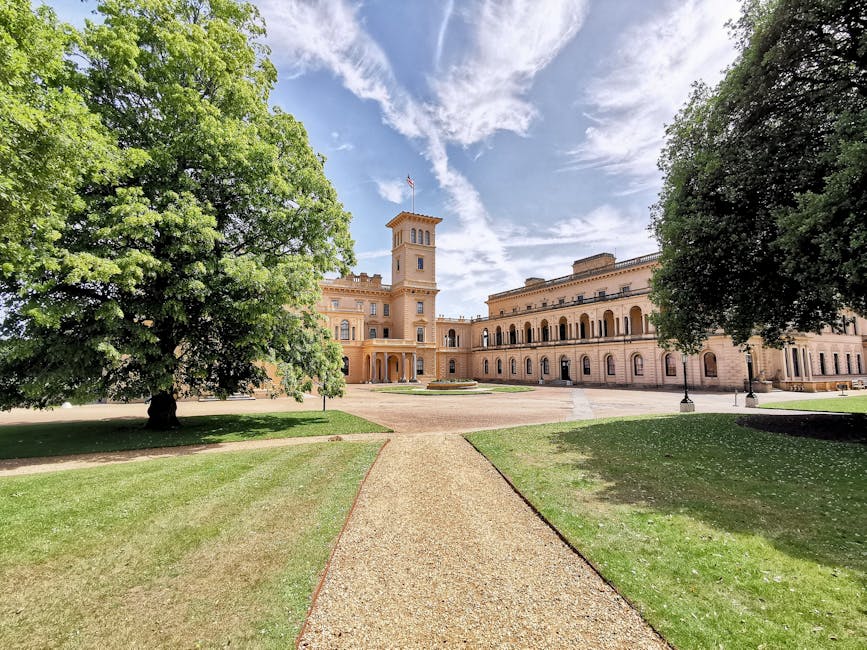A clear view of Syon Park's historic building, featuring a tall central tower and arched windows, painted in a warm beige tone. In the foreground, a gravel pathway leads directly to the building's entrance, flanked by well-maintained lawns and large, leafy green trees on both sides. The scene is lit by natural daylight under a partly cloudy sky, creating a bright and welcoming atmosphere. This outdoor setting is associated with house removals and relocation services, as the image reflects the setting where professional movers like Man and Van Isleworth may load or unload furniture and packing materials during a home relocation process near this landmark.