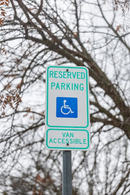 A vertical metal parking sign mounted on a pole with a background of leafless tree branches and an overcast sky. The sign has a white background with teal borders and teal text reading 'RESERVED PARKING' at the top, a blue square with a white wheelchair symbol in the center, and teal text below that states 'VAN ACCESSIBLE'. This signage indicates designated parking, including access for disabled vehicles, and is situated near an area where house removals or furniture transport may occur. Man and Van Isleworth, a professional removals company, uses such signage to facilitate smooth moving logistics, particularly when navigating narrow access points or obtaining necessary permits for home relocation projects, as detailed on their Syon Park home removals guide page.