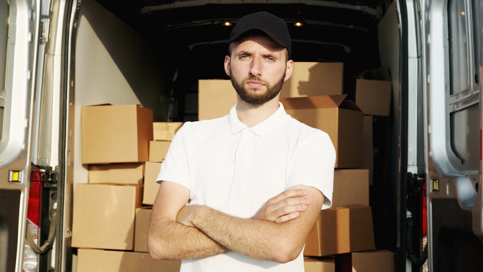 A young male mover with a serious expression, wearing a black cap and a white polo shirt, stands inside the open rear of a moving van. The van's interior is filled with neatly stacked cardboard boxes of various sizes, some wrapped in plastic or packing materials. The boxes are arranged to maximize space and ease loading and unloading. The scene is set outdoors on a paved surface, with natural light illuminating the area. Visible are the metal sides of the van, with some safety lights and reflectors. The mover crosses his arms in front of his chest, indicating readiness for the furniture transport or home relocation process, which is consistent with professional removals services provided by Man and Van Isleworth. This image captures the preparation and loading stage of packing and moving, highlighting efficient packing and handling during a house removal or furniture transport service.