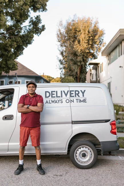A young male mover with a serious expression, wearing a black cap and a white polo shirt, stands inside the open rear of a moving van. The van's interior is filled with neatly stacked cardboard boxes of various sizes, some wrapped in plastic or packing materials. The boxes are arranged to maximize space and ease loading and unloading. The scene is set outdoors on a paved surface, with natural light illuminating the area. Visible are the metal sides of the van, with some safety lights and reflectors. The mover crosses his arms in front of his chest, indicating readiness for the furniture transport or home relocation process, which is consistent with professional removals services provided by Man and Van Isleworth. This image captures the preparation and loading stage of packing and moving, highlighting efficient packing and handling during a house removal or furniture transport service.